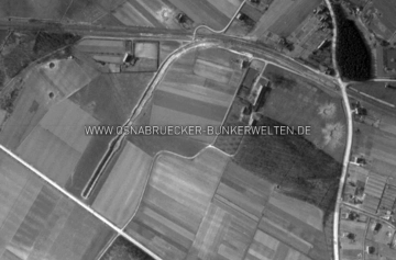 Eine Luftaufnahme des Zweiggleises am Sutthauser Bahnhof, aufgenommen im März 1945. Deutlich sind hier die Erdaufwürfe seitlich des Gleises zu erkennen. Bei genauerer Betrachtung sind zudem drei bzw. vier Ausbuchtungen zu erkennen, die auf die Stationierung von Geschützwagen einer Eisenbahnflak hindeuten könnten. Zur Orientierung: Unten links verläuft die Malberger Staße, rechts von oben nach unten die heutige Hermann-Ehlers-Straße, die das Hauptgleis auf Höhe des Sutthauser Bahnhofs kreuzt. Mittig am oberen Bildrand ist ein Teil der Straße Am Knochenhof zu erkennen. Luftaufnahme Zweiggleis Bahnhof Sutthausen
