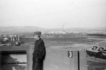 In der Flak-Stellung auf dem Westerberg in Osnabrück (Rückseitiger Text: "Blick auf die Stadt"). Hinter der Wiese sind die Baracken der Metzer- und Scharnhorstkaserne zu erkennen. Dahinter der Hafen und die Speicher der Winkelhausen-Kaserne. Im Hintergrund am linken Rand der Piesberg. Wann und durch wen die Aufnahme entstand, ist unklar. Auch der Name des abgebildeten Soldaten wurde nicht vermerkt. Flak-Stellung Westerberg, Blick auf Kasernen und Hafen